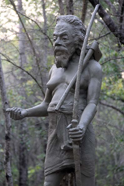 Outdoor cement statue of chieftain Thisa Hamy holding axe and walking staff in forest sanctuary